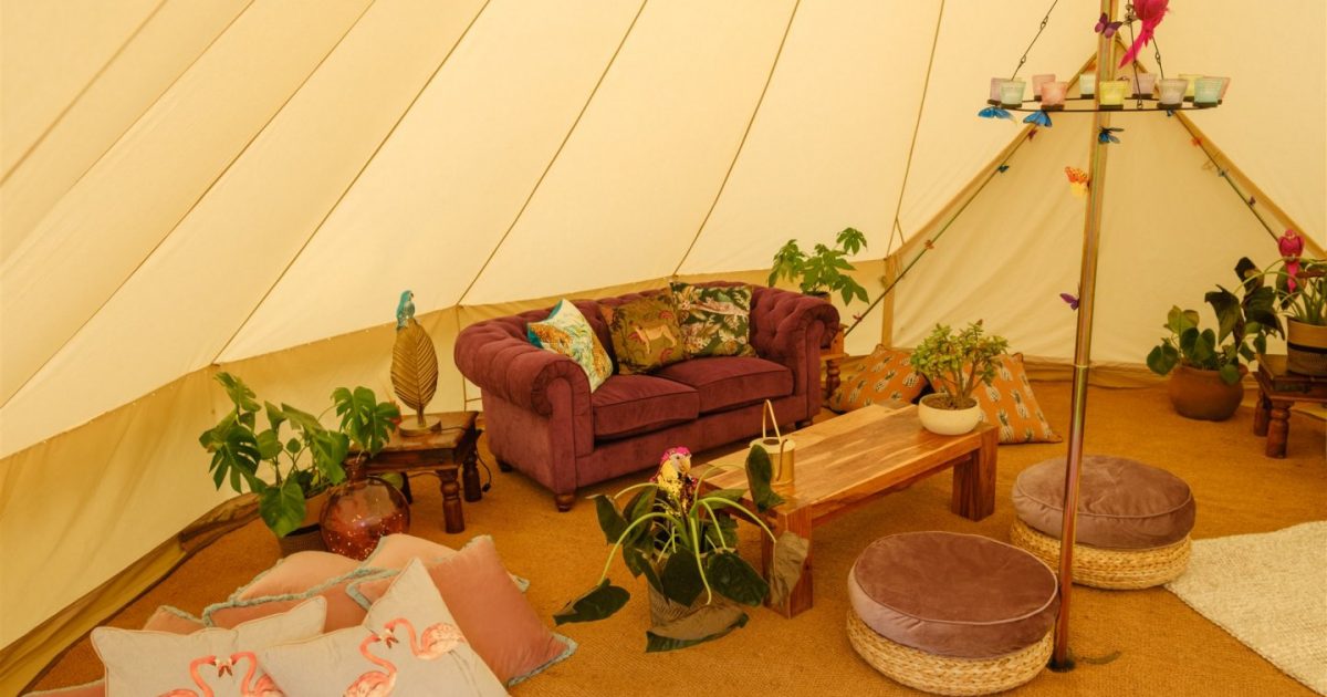 Interior of 7m chill out bell tent at hen camp. Decorated with purple sofa, tropical cushions, and plants for hen party glamping.